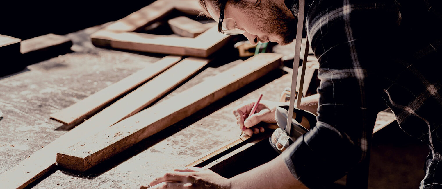 Ein Tischler zeichnet konzentriert Maße auf einem Brett mit einem Gliedermaßstab und einen Bleistift ein. Er beugt sich über die Werkbank. Im Hintergrund sind weiter Holzzuschnitte und die Werkstatt zu erkennen. 