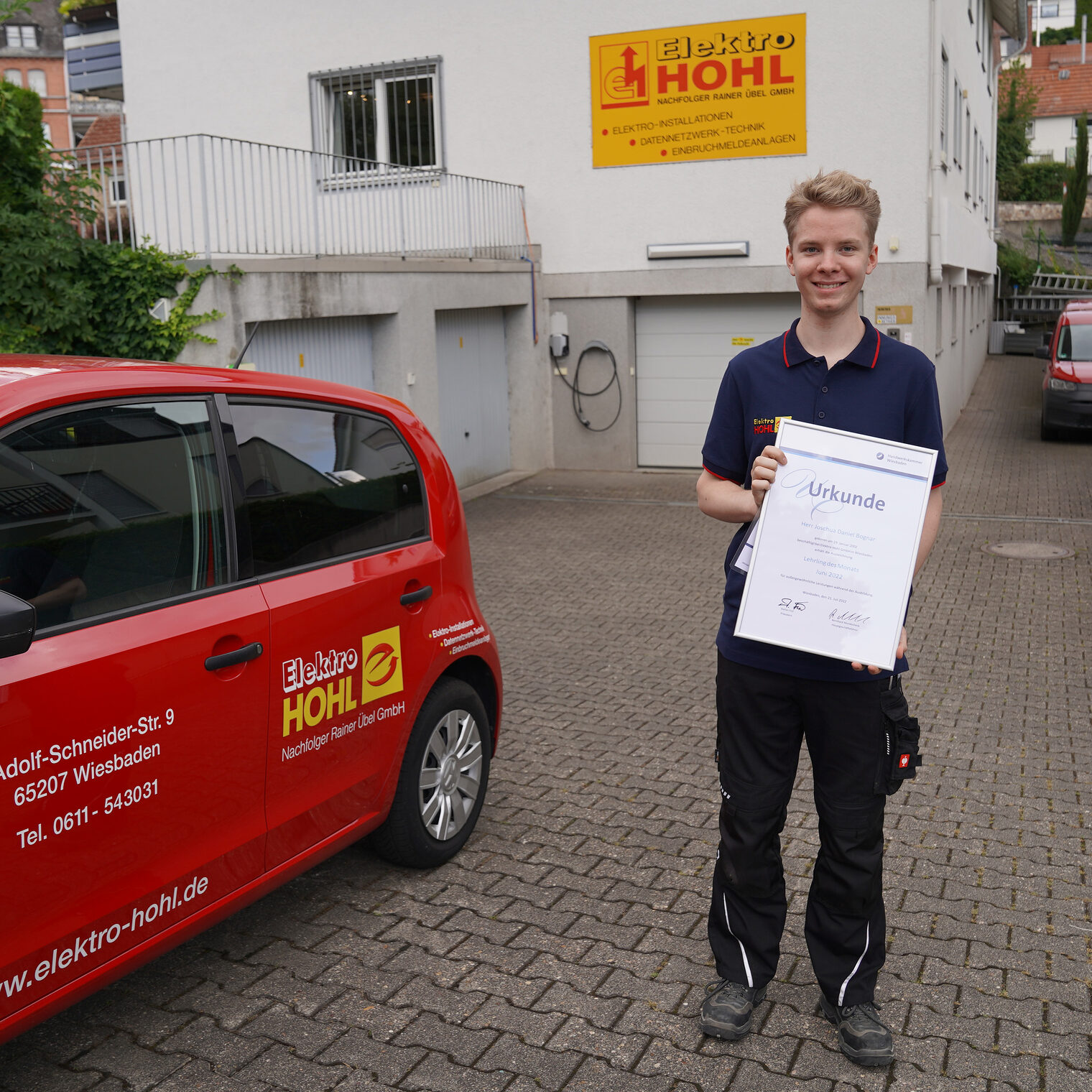 Junger Mann hält eine Urkunde vor sich. Er steht in einer Hofeinfahrt neben einem Firmen-PKW.