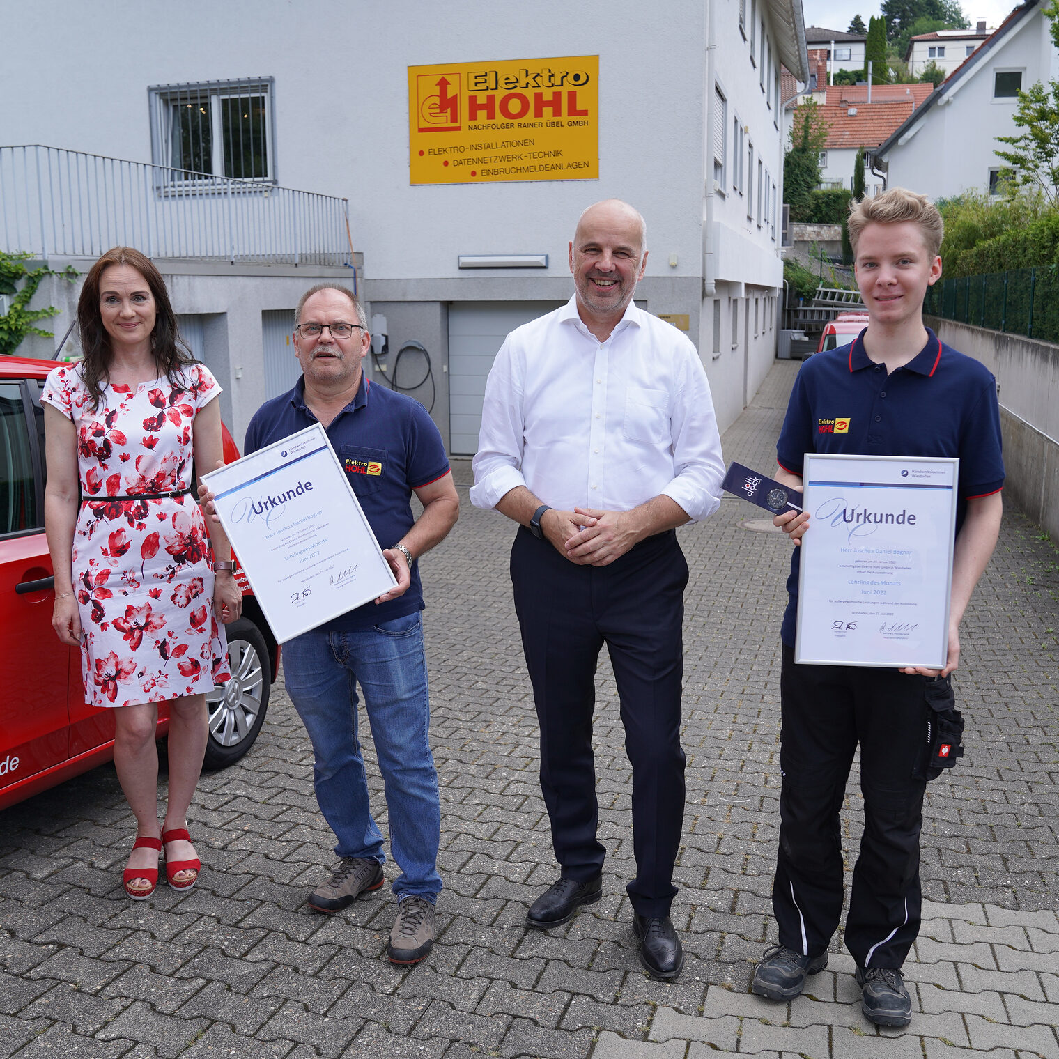 Vier Personen stehen neben Firmen-PKW in einer Hofeinfahrt. Junger Mann rechts 2. v. links halten eine Urkunde.