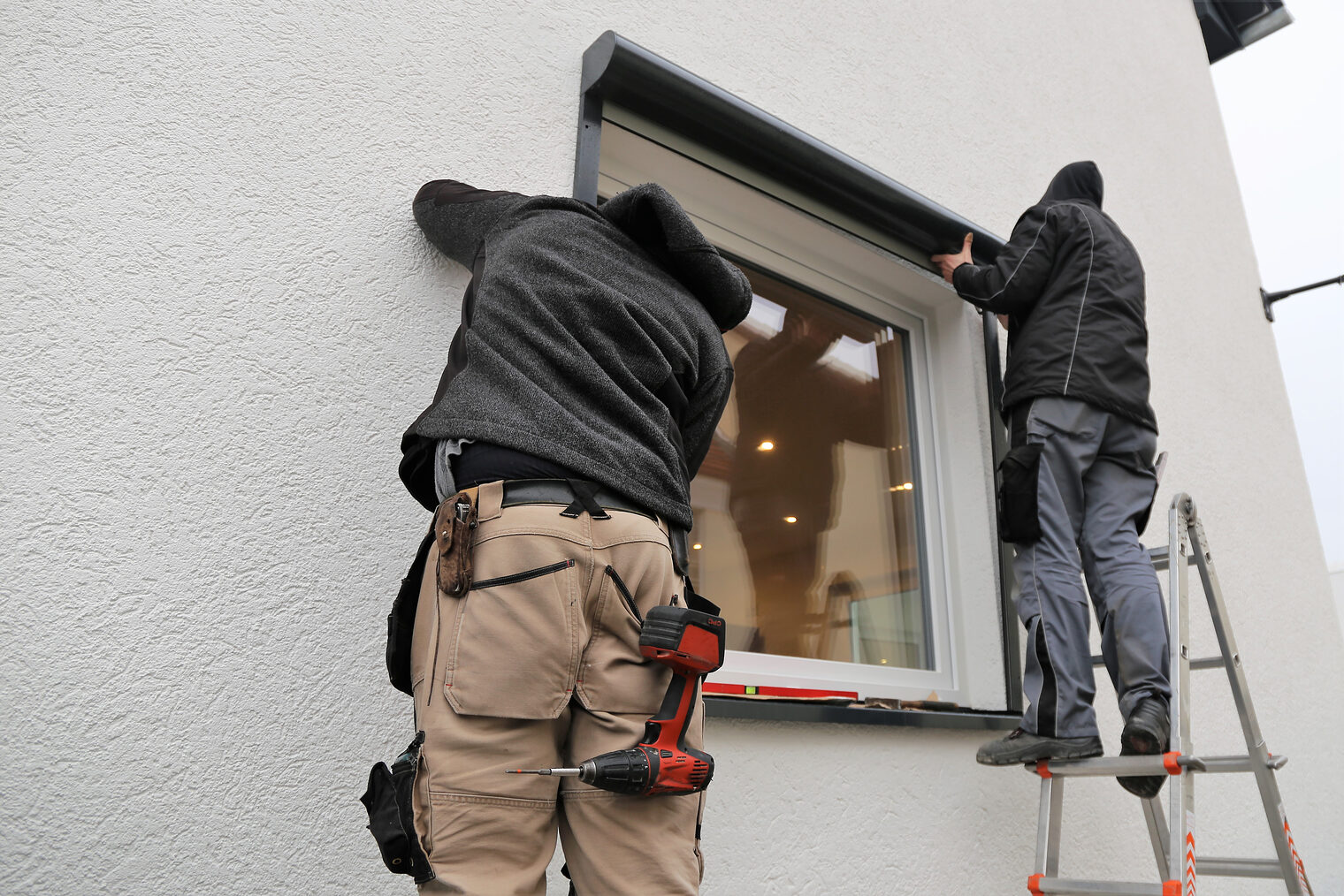 Zwei Arbeiter montieren einen Rolladenkasten an eine Hausfassade. 