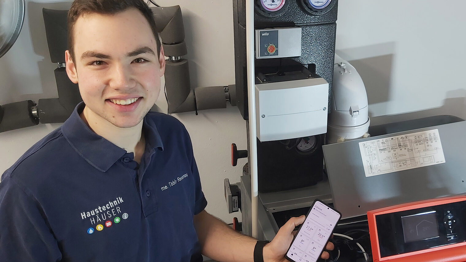 Tobin Jonas Rosenau, Meister im Installateur- und Heizungsbauerhandwerk, steht an einer Heizungsanlage mit einem Handy in der Hand, um die Digitalisierung in seinem Handwerk zu demonstrieren.