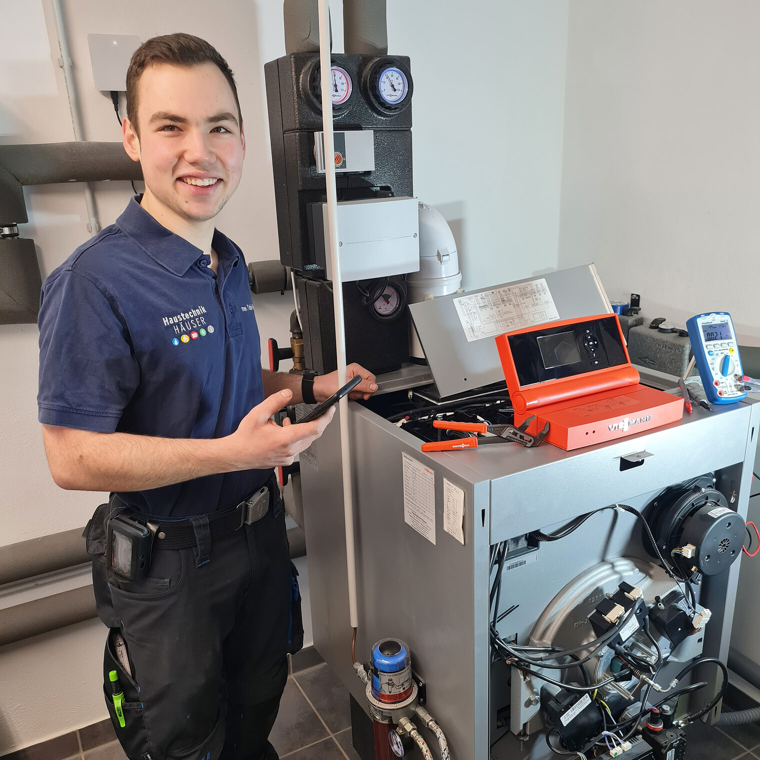 Tobin Jonas Rosenau, Meister im Installateur- und Heizungsbauerhandwerk, steht an einer Heizungsanlage mit einem Handy in der Hand, um die Digitalisierung in seinem Handwerk zu demonstrieren.