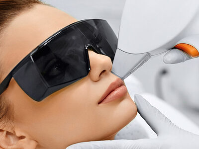 Frauenkopf mit dunkler Brille liegt auf weißer Liege. Zwei Hände mit weißen Handschuhen halten ein Lasergerät an die Wange.