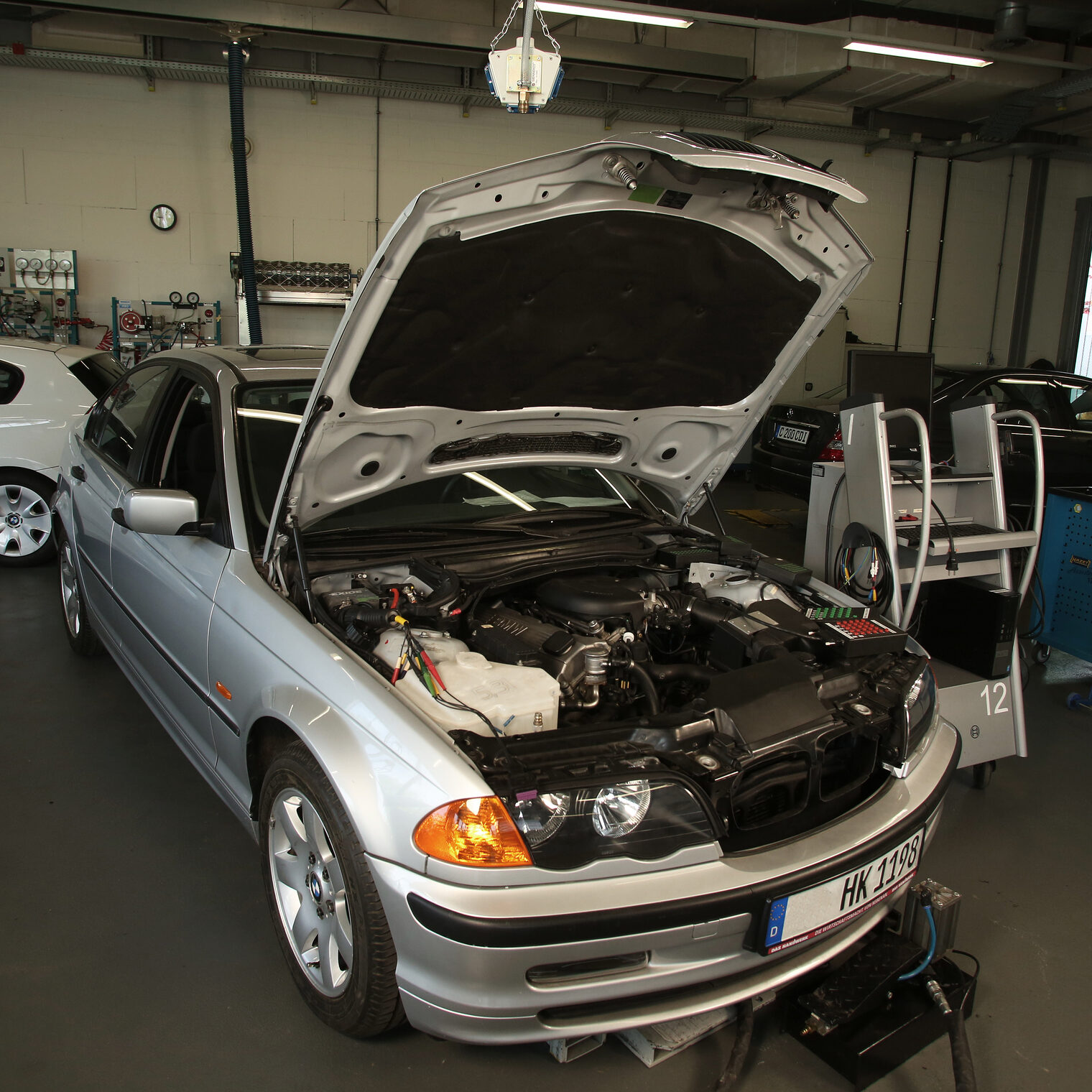Ein silbernes Auto mit offener Motorhaube steht in einer Werkstatt. Ein Analysegerät steht rechts neben dem Auto. Weiter Autos sind im Hintergrund zu sehen. 