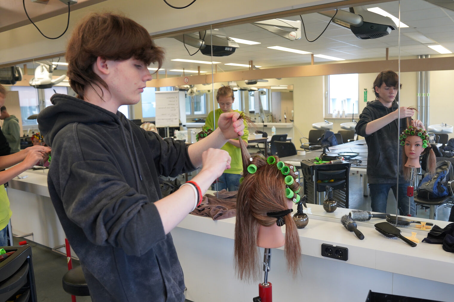 Ein junger Schüler bei einer Berufsorientierung in der Friseur-Lehrwerkstatt, der Lockenwickler auf einem Übungskopf eindreht. 