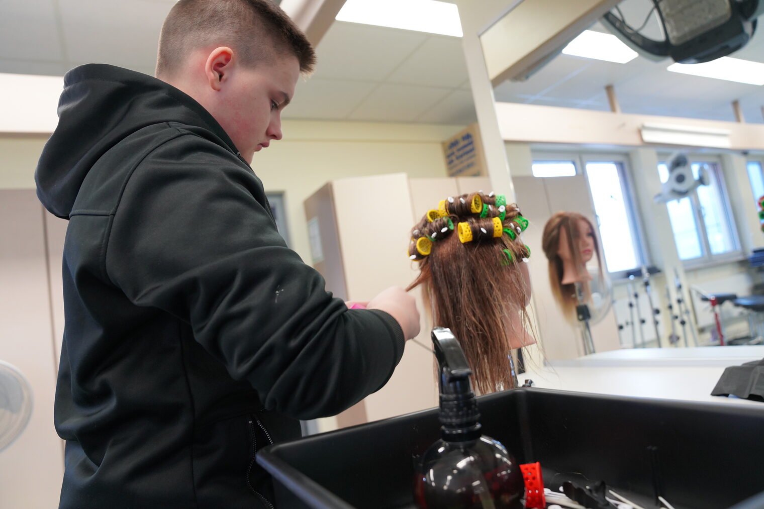 Ein junger Schüler bearbeitet während der Berufsorientierungswoche einen Friseurübungskopf. Die Aufgabe ist Lockenwickler einzudrehen. 