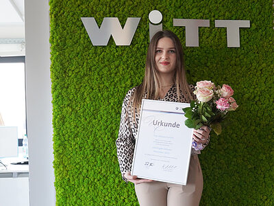Eine junge Frau steht in einem Büroraum vor einer grünen Wand und hält eine Urkunde und einen Blumenstrauß vor sich.