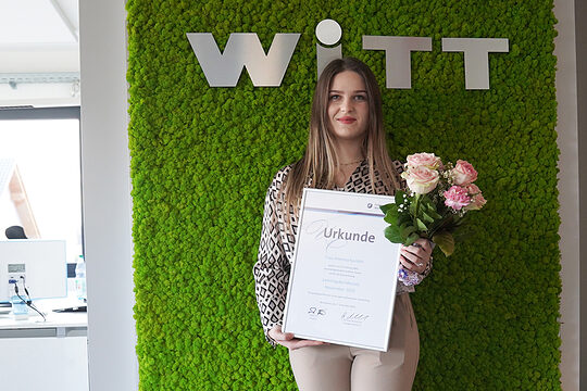 Eine junge Frau steht in einem Büroraum vor einer grünen Wand und hält eine Urkunde und einen Blumenstrauß vor sich. 