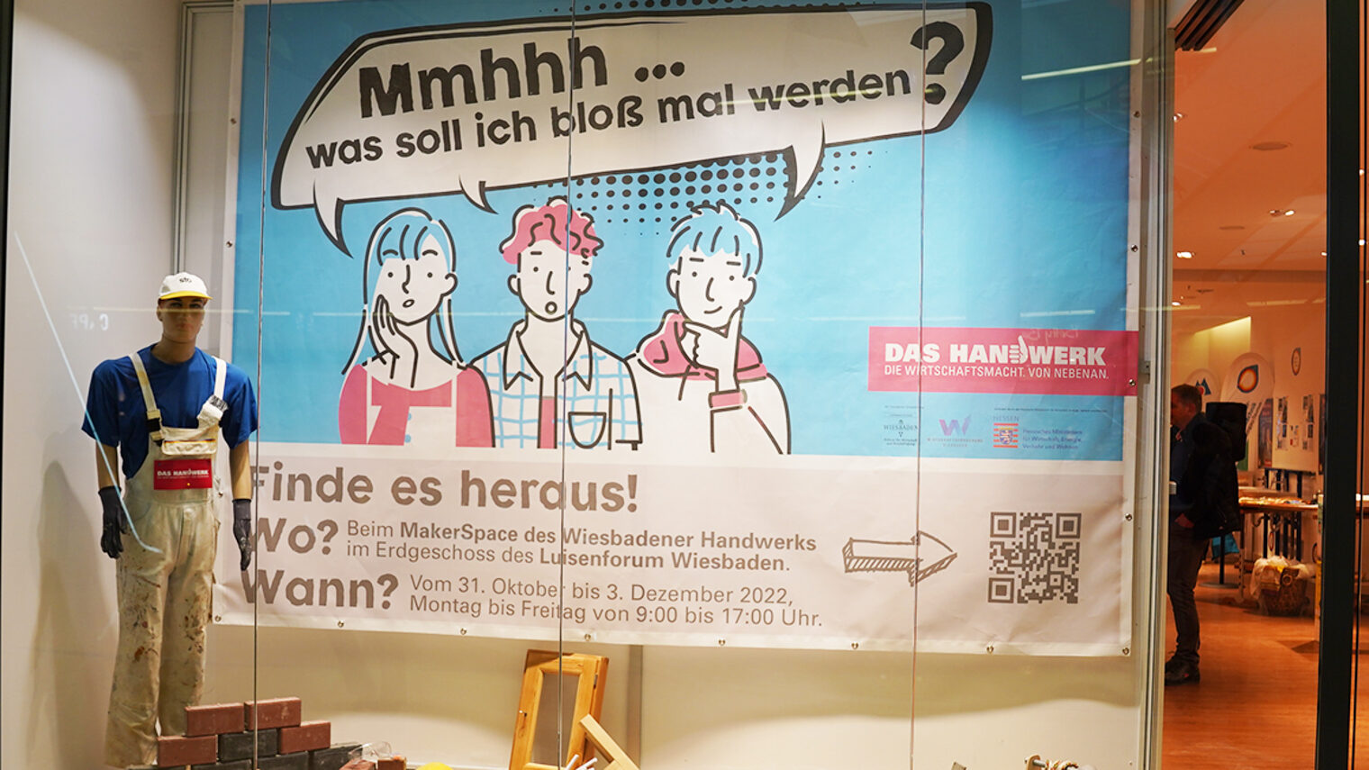 Ansicht eines Schaufensters. Links steht eine Schaufensterpuppe in weißer Latzhose als Handwerker angezogen. in der Mitte hängt ein Plakat auf dem in Comic-Zeichnung 3 junge Leute sind. Über ihnen steht in einer Sprechblase "Mmhhh... was soll ich bloß mal werden?"