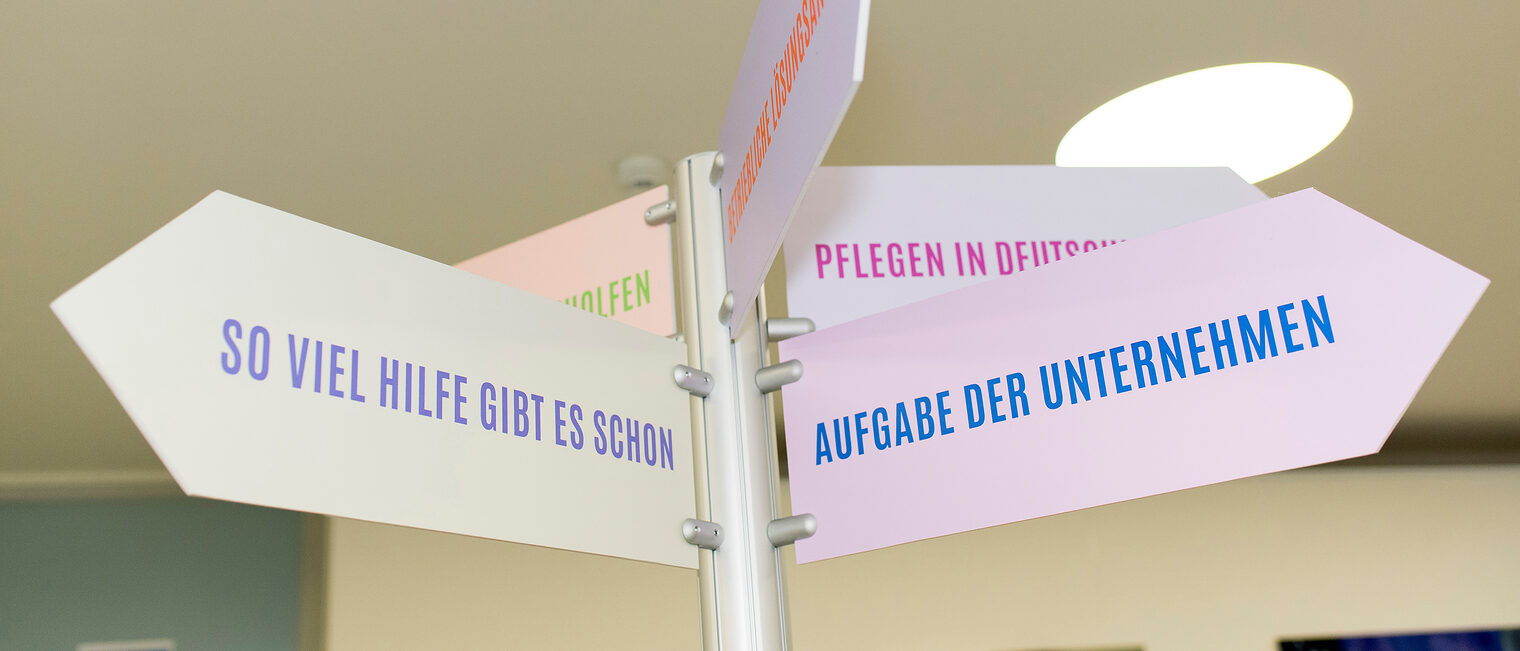 Pflege und Beruf - was bedeutet das für Arbeitgeber? Richtungspfeile mit verschiedenen Aufschriften wie: So viel Hilfe gibt es schon; Aufgabe der Unternehmen. Diese Richtungspfeile sind Teil einer Wanderausstellung.