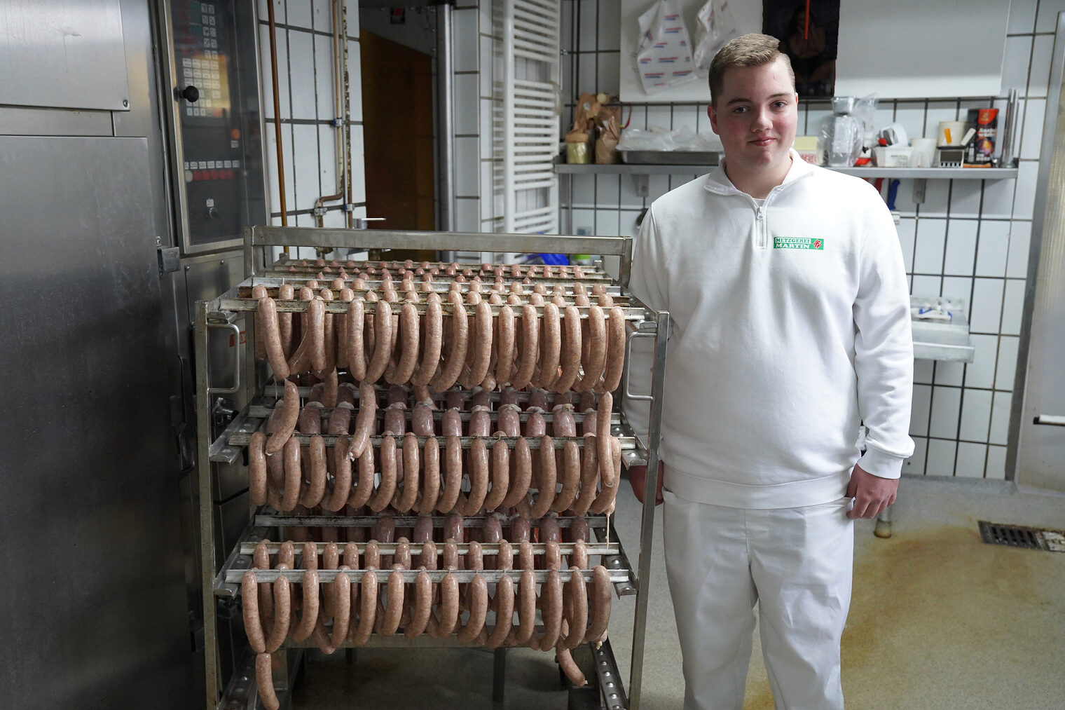 Frontalaufnahme eines jungen Mannes in weißer Fleischer-Arbeitskleidung, neben einem Metallgestell mit frischen Würsten.