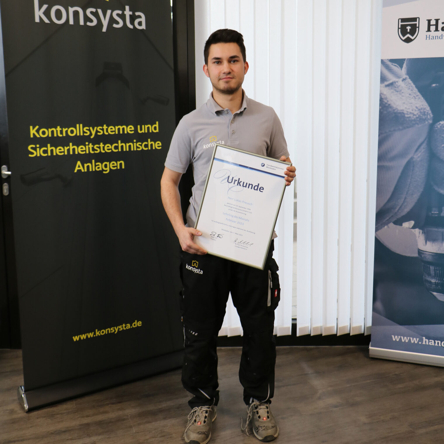 Junger Mann steht vor einem Banner der Firma Konsysta GmbH. vor sich hält er eine Urkunde zum "Lehrling des Monats".