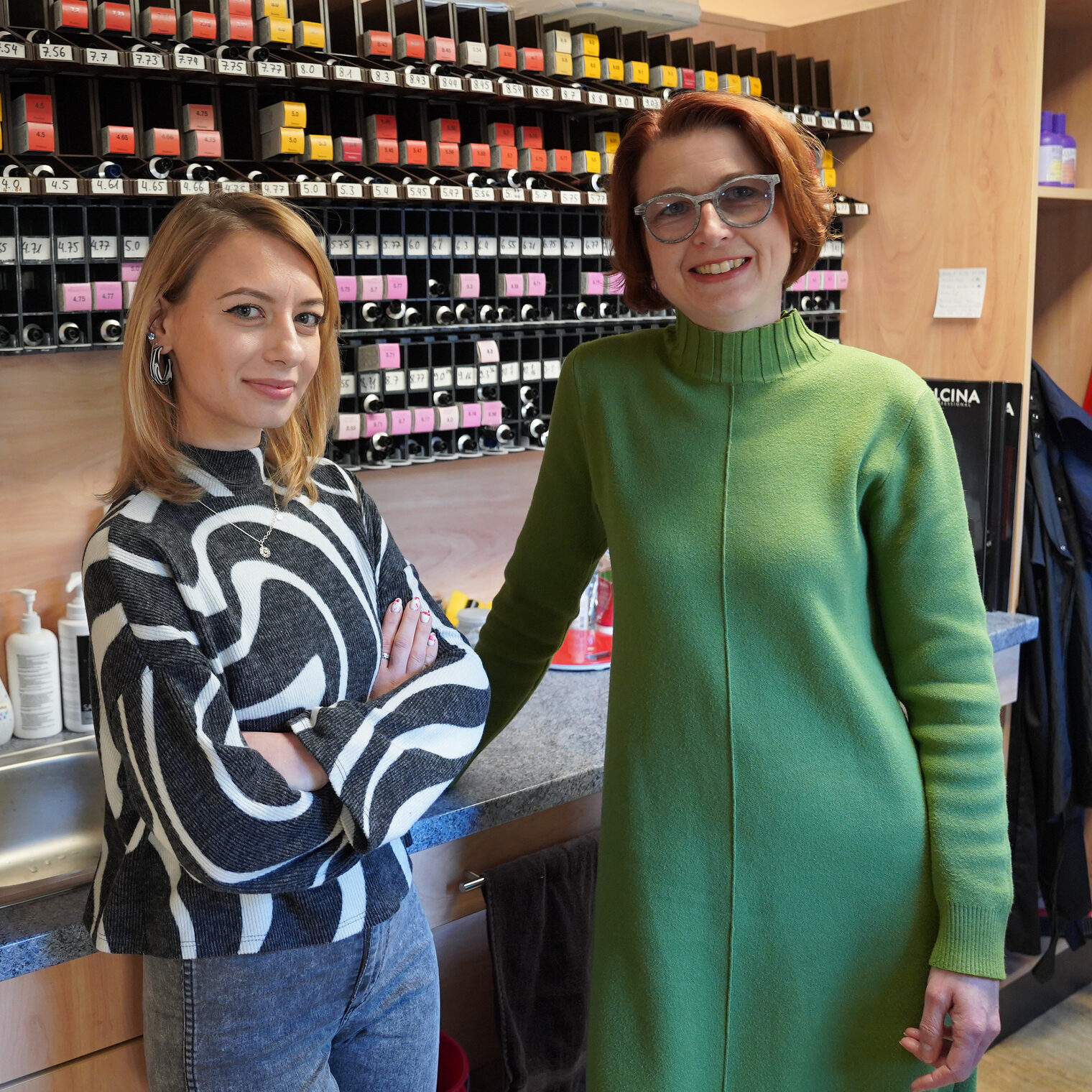 Pia Henrich und Daria Karlova sind auf dem Bild im Friseursalon von Pia Henrich zu sehen. Beide stehen vor dem Regal, indem die Colorationsfarben aufbewahrt werden. 
