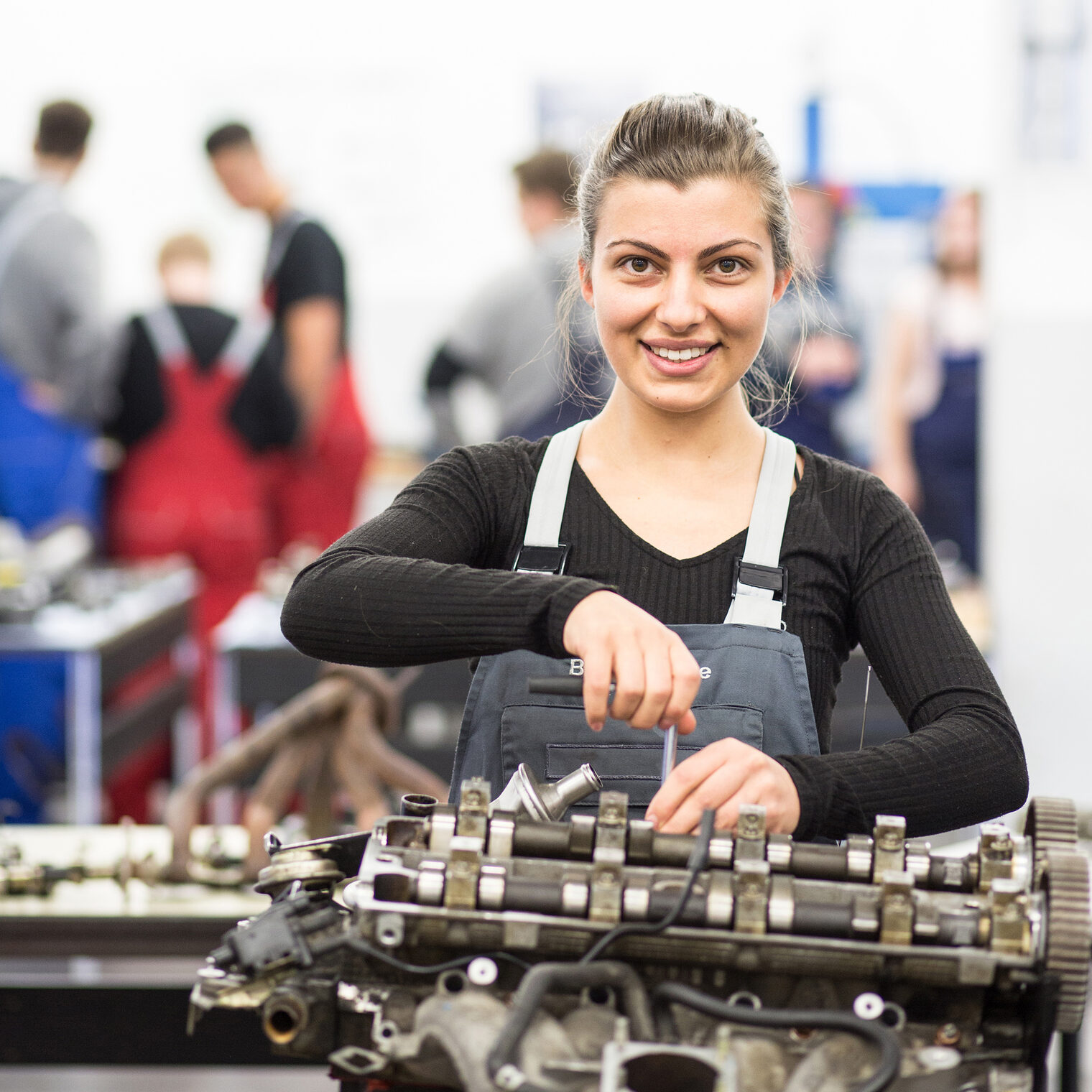 Eine junge Frau, Kraftfahrzeugtechnikerin, lächelt in die Kamera und steht vor einem Motorblock und hat ein Werkzeug in der Hand.Schlagwort(e): Berufsbildung, Bildung, Bildungszentrum, Braunschweig, Meisterkurs, Seminare, TZH, Technologiezentren, Technologiezentrum, Weiterbildung, fortbildung, kfz, meister, seminar, unterricht, ÜLU, Überbetriebliche Lehrlingsunterweisung