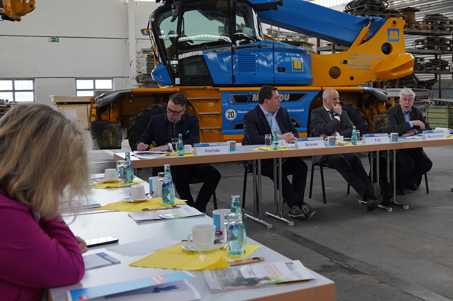 Teilnehmende des Pressegesprächs sitzen an Tischen vor einem Baukran in einer Bauhalle.