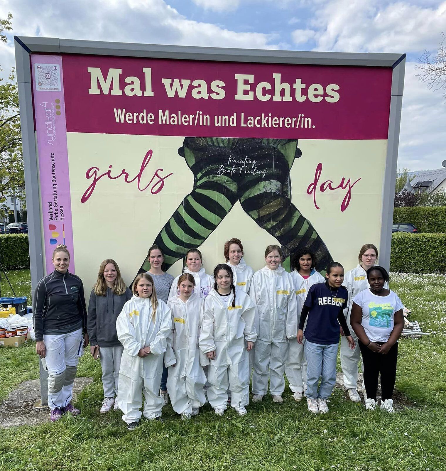 Eine junge Frau und eine Gruppe jugendlicher Mädchen in weißen Maleranzügen stehen vor einem Großflächenplakat mit der Aufschrift "Girls¿day".