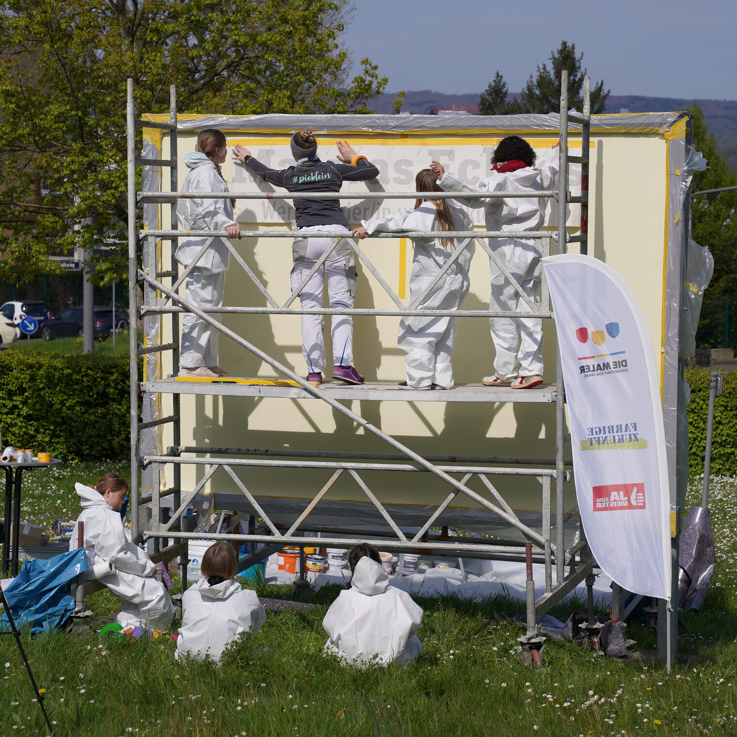 Jugendliche Mädchen in weißen Maleranzügen stehen auf einem Gerüst am Großflächenplakat der Handwerkskammer Wiesbaden und bemalen es.