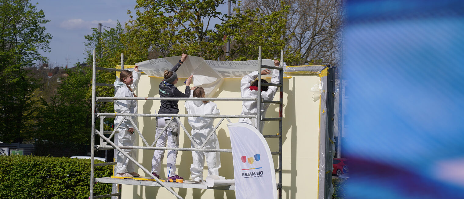 Malermeisterin Jessica Jörges und eine kleine Gruppe Mädchen in Maleranzügen bereiten Farbe und Werkzeug für Malerarbeiten vor.