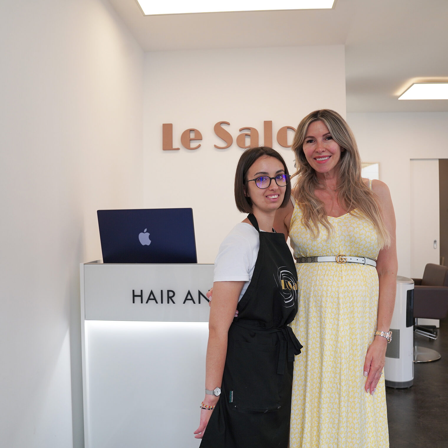 Marie Dombre lernte im Friseursalon Le Salon Bisera in Wiesbaden den Arbeitsalltag von Friseurmeisterin Bisera Ukikj (rechts) kennen. Das Bild zeigt die beiden Damen Arm in Arm im Friseursalon vor dem Empfangstresen. 