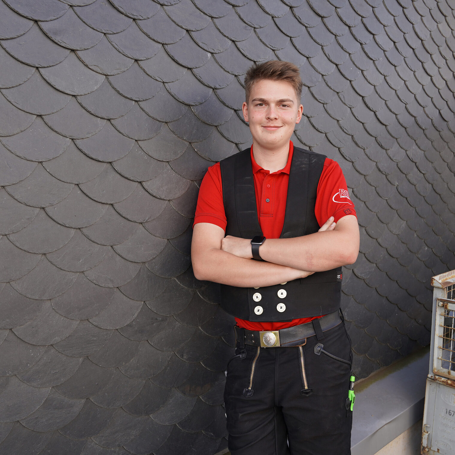 Das Bild zeigt Leon Hain wie er entspannt mit verschränkten Armen vor einer Schieferwand im Berufsbildungswerk des Hessischen Dachdeckerhandwerks in Weilburg steht und in die Kamera lächelt.