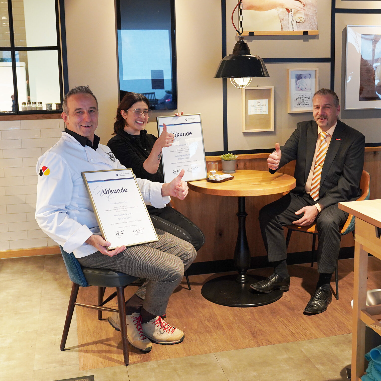 Bäckermeister Braun und Lehrling des Monats Bonita Fischer mit Urkunden sowie Kammervizepräsident Andreas Brieske sitzen am Tisch im Café.