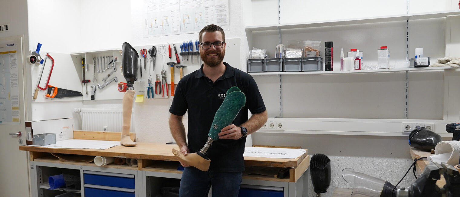 Jan Thorben Balschun steht in seiner Werkstatt, lächelt in die Kamera und hält eine Unterschenkelprothese in der Hand.