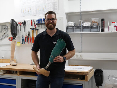 Jan Thorben Balschun steht in seiner Werkstatt, lächelt in die Kamera und hält eine Unterschenkelprothese in der Hand.
