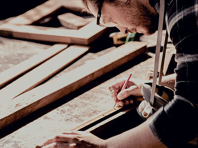 Ein Tischler zeichnet konzentriert Maße auf einem Brett mit einem Gliedermaßstab und einen Bleistift ein. Er beugt sich über die Werkbank. Im Hintergrund sind weiter Holzzuschnitte und die Werkstatt zu erkennen.
