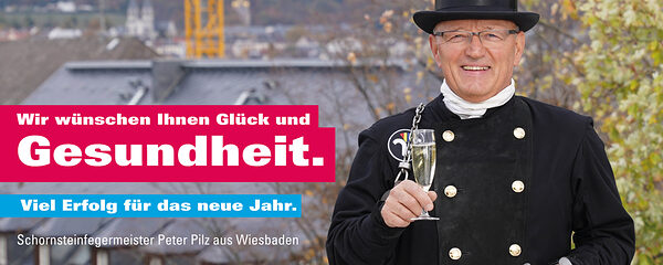 Schornsteinfegermeister Peter Pilz auf dem Dach mit einem Glas Sekt und einem Hufeisen in der Hand und wünscht somit dem Betrachter Glück und Gesundheit für das neue Jahr.