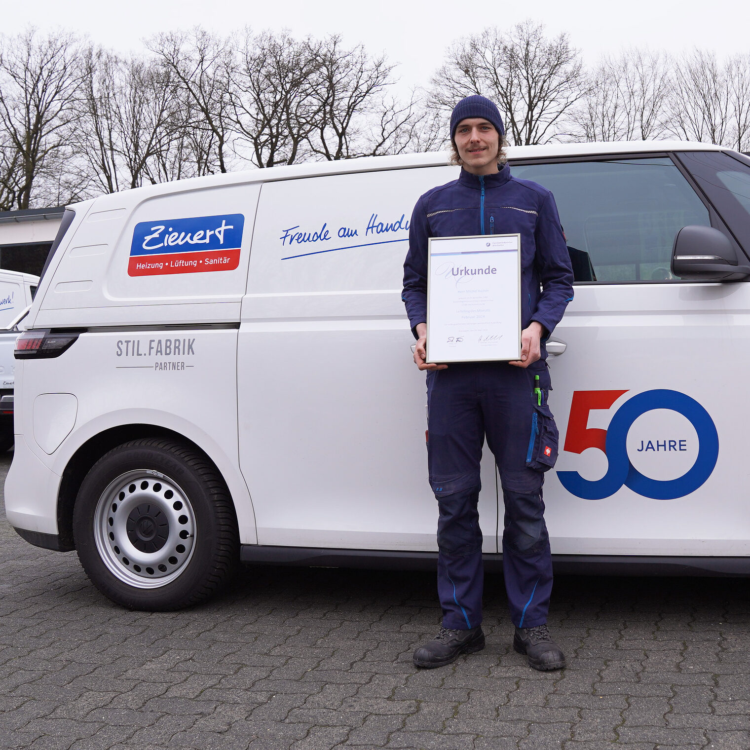 Lehrling des Monats Februar 2024 steht vor einem Firmenwagen der Fa. Zienert GmbH aus Sinn und hält seine Urkunde in der Hand.