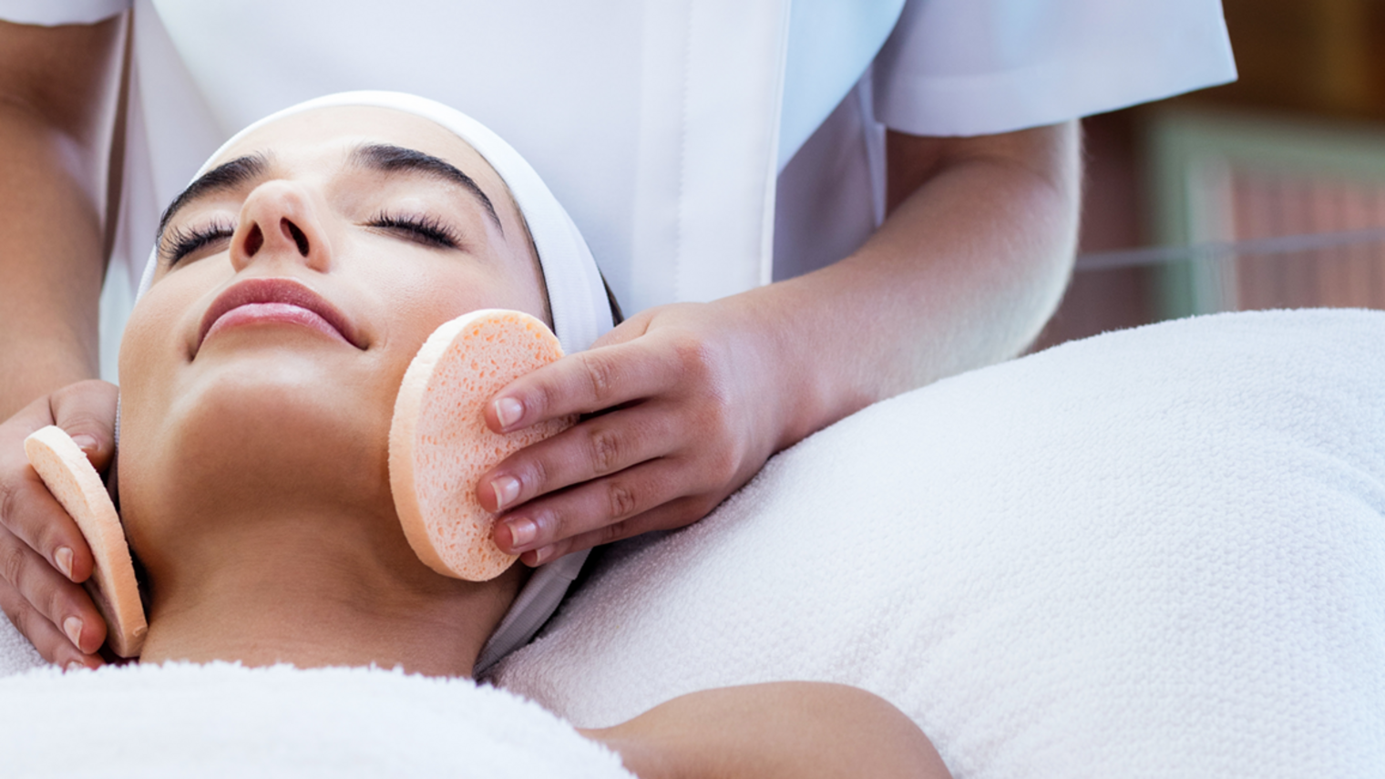 Das Bild zeigt eine Kosmetikerin, vor ihr die Kundin, der sie gerade das Gesicht mit Schwämmen reinigt. 