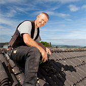 Dachdecker sitzt bei Sonnenschein auf dem Dach, blauer Himmel im Hintergrund und lächelt in die Kamera. 
