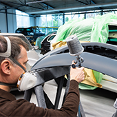 Bild zeigt einen Fahrzeuglackierer mit einer Lackierpistole ein Autoteil besprühen. Dabei trägt er eine Maske. 