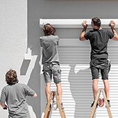 Drei Rollladen- und Sonnenschutzmechatroniker montieren an einem Haus eine Jalousie an. 
