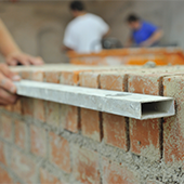 Das Bild zeigt eine Mauer in Arbeit, eine Hand, die ein Blech hält um die Mauer zu überprüfen, ob diese gerade ist. Im Hintergrund sind verschwommen zwei Menschen zu sehen. 