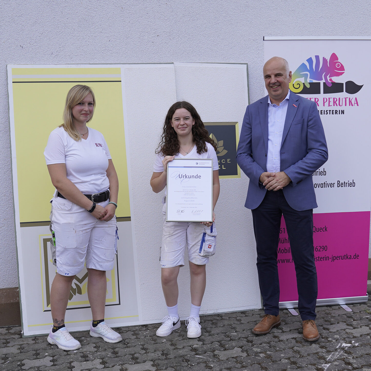von links: Meisterin im Maler und Lackierer-Handwerk Jennifer Perutka, Lehrling des Monats Lea Semmler und Kammerpräsident Stefan Füll, stehend vor einer gestalteten Wandtafel.