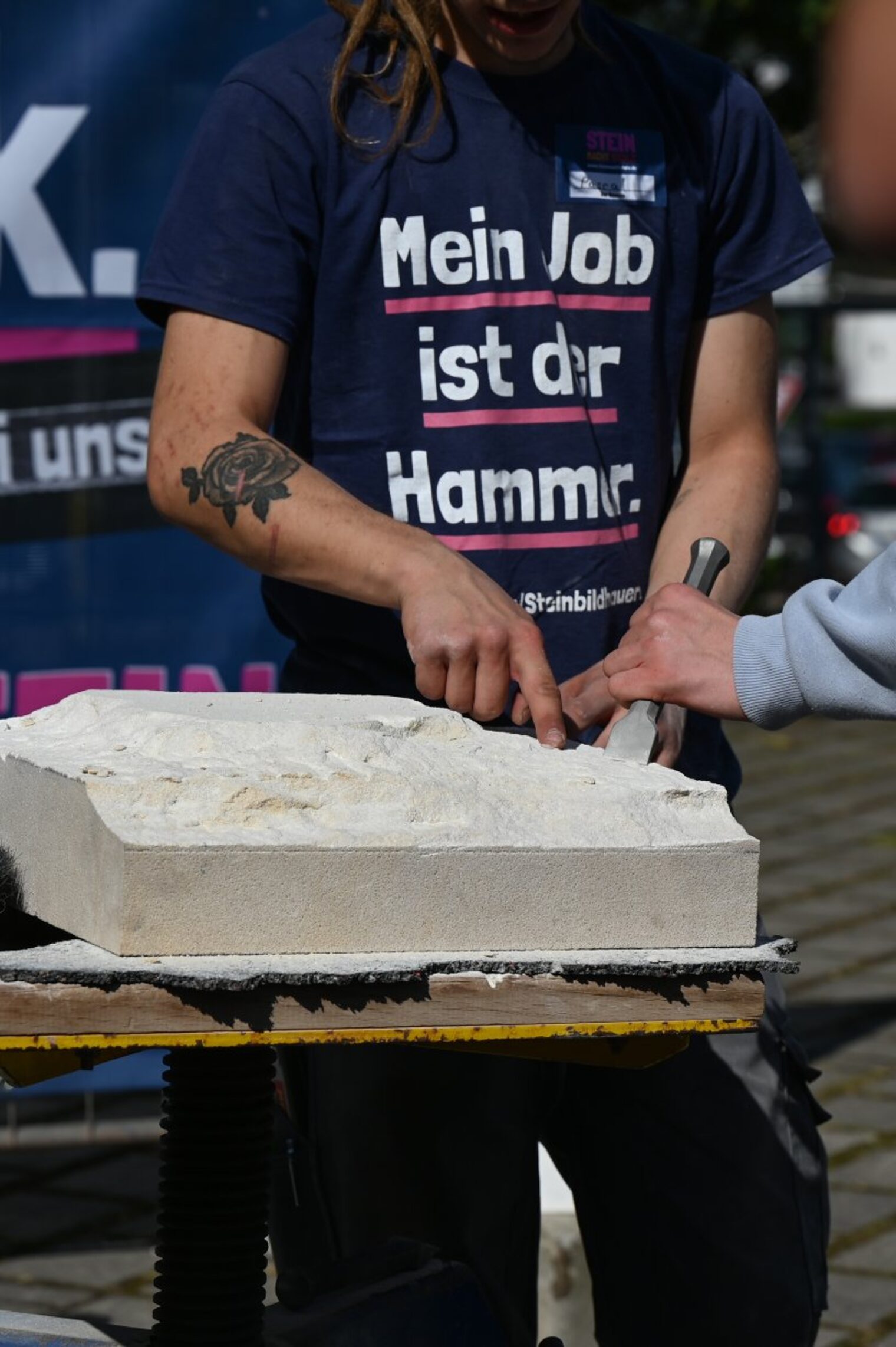Auf dem Bild sieht man einen zu bearbeitenden Stein am Stand der Steinmetze. Es ist auch eine bearbeitende Hand zu sehen, sowie eine helfende Person. 