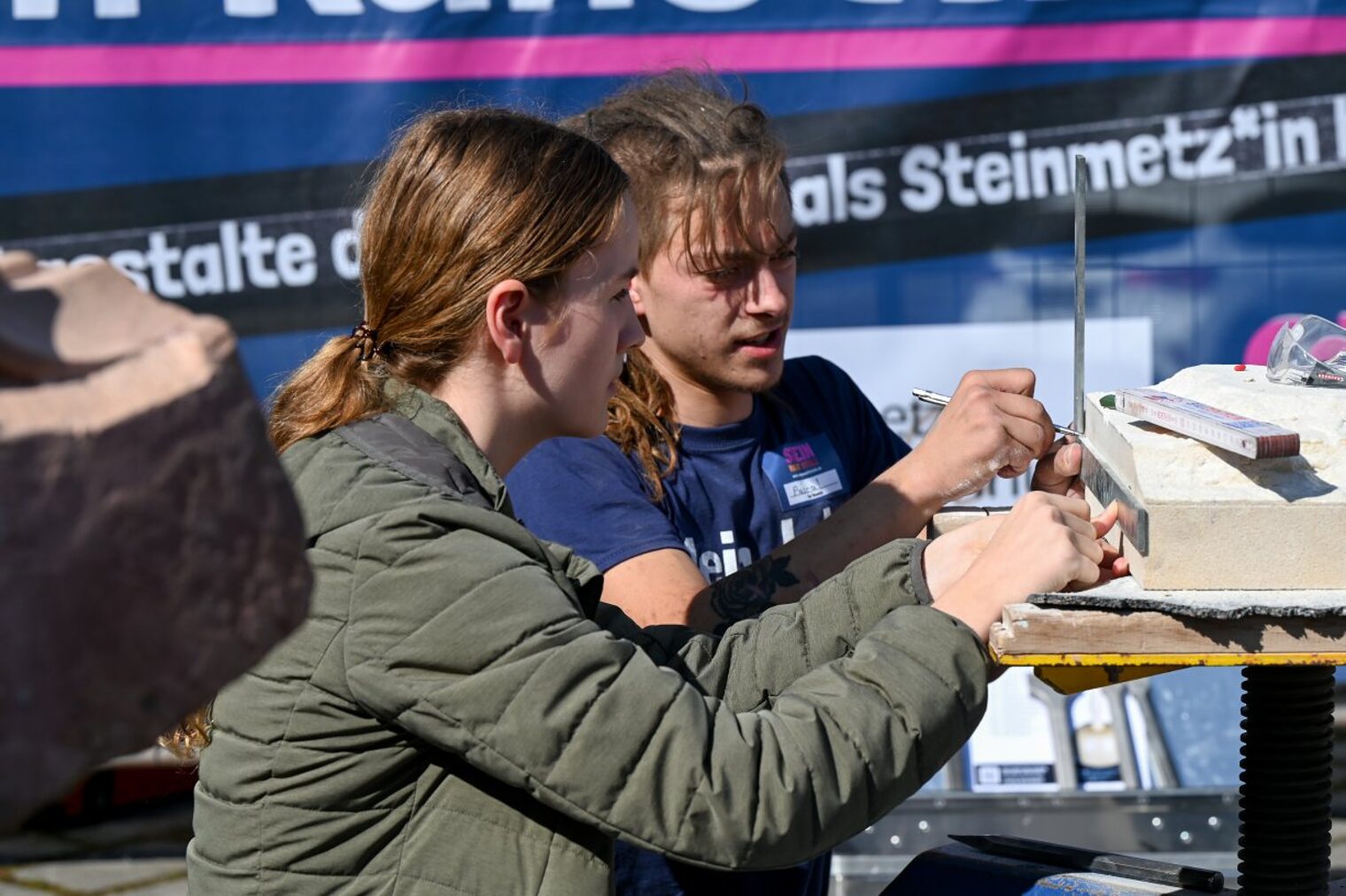 Ein Steinmetz zeigt einer interessierten jungen Frau, wie man einen Stein bearbeitet. 