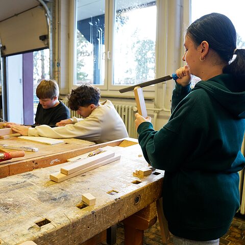 Es sind drei Schüler der Theodor-Fliedner-Schule zu sehen während Sie an einem Holzprojet für Liegestühle arbeiten und dazugehörige Holzteile entsprechen schleifen und formen.