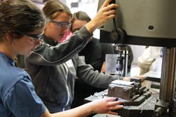 Zwei Schülerinnen die unter Anleitung eine Ständebohrmaschine in einer Werkstatt bedienen.
