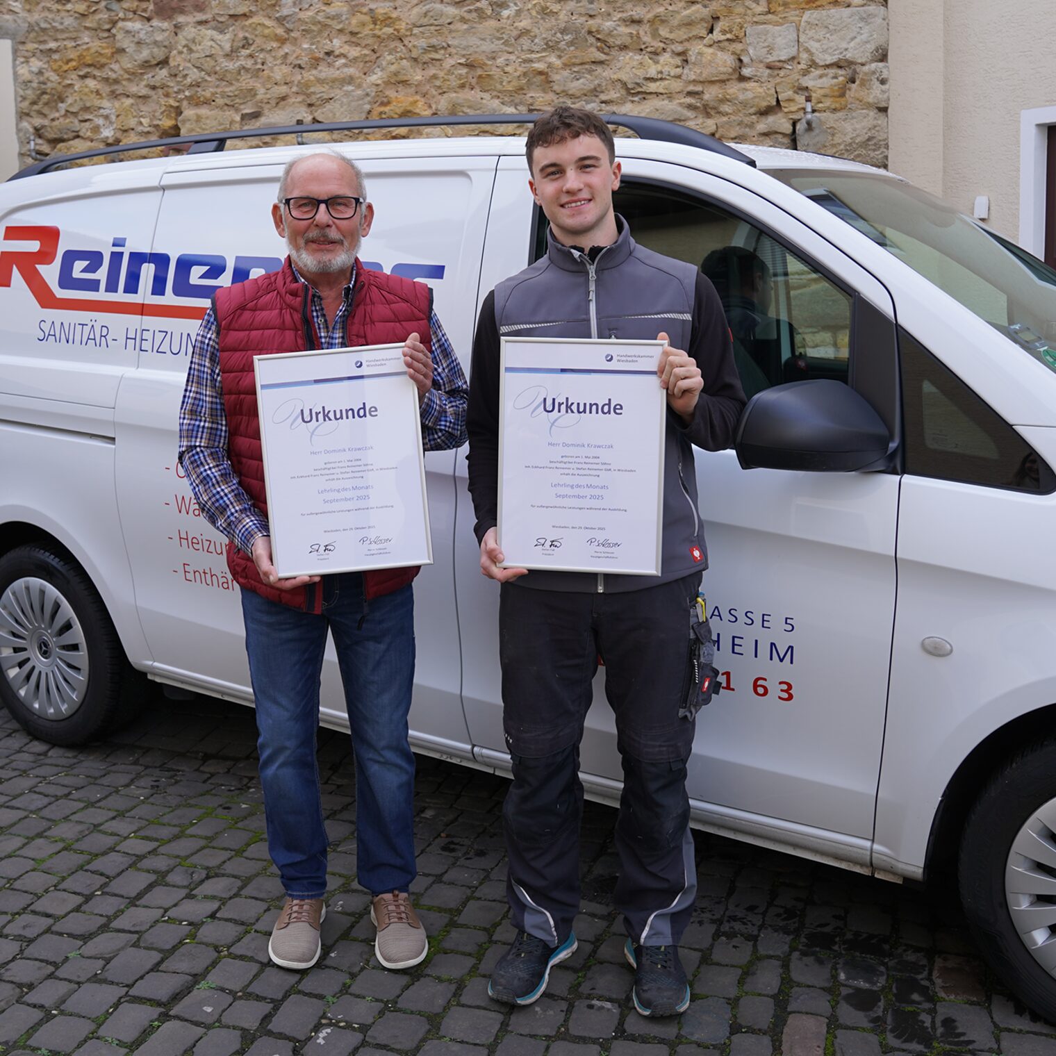 Dominik Krawczak und Ausbilder Eckhard Reinemer mit den Urkunden vor einem Firmenfahrzeug.