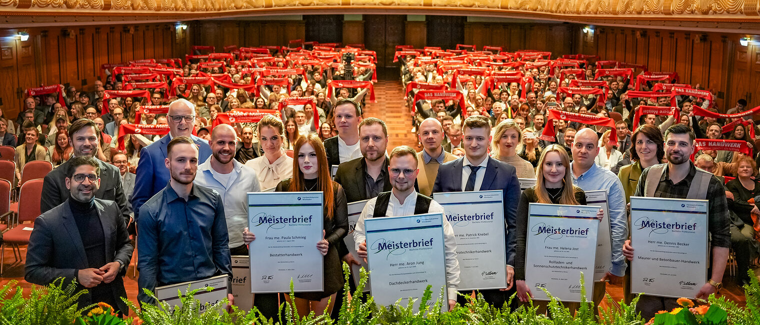 Gruppenfoto der Jahresbesten Jungmeisterinnen und Jungmeister mit Ihrem Meisterbrief mit Blick auf den Saal.