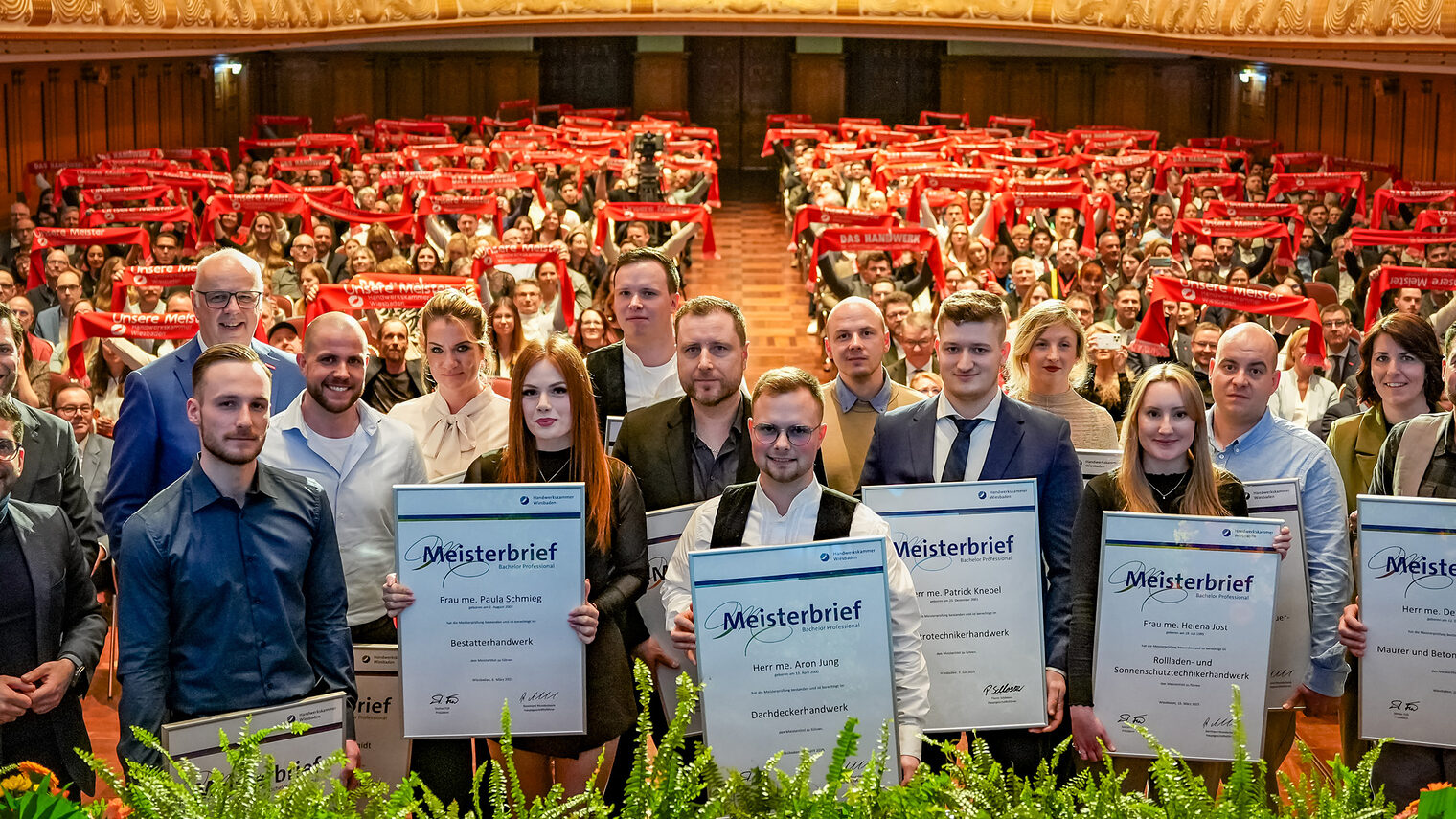 Gruppenfoto der Jahresbesten Jungmeisterinnen und Jungmeister mit Ihrem Meisterbrief mit Blick auf den Saal.