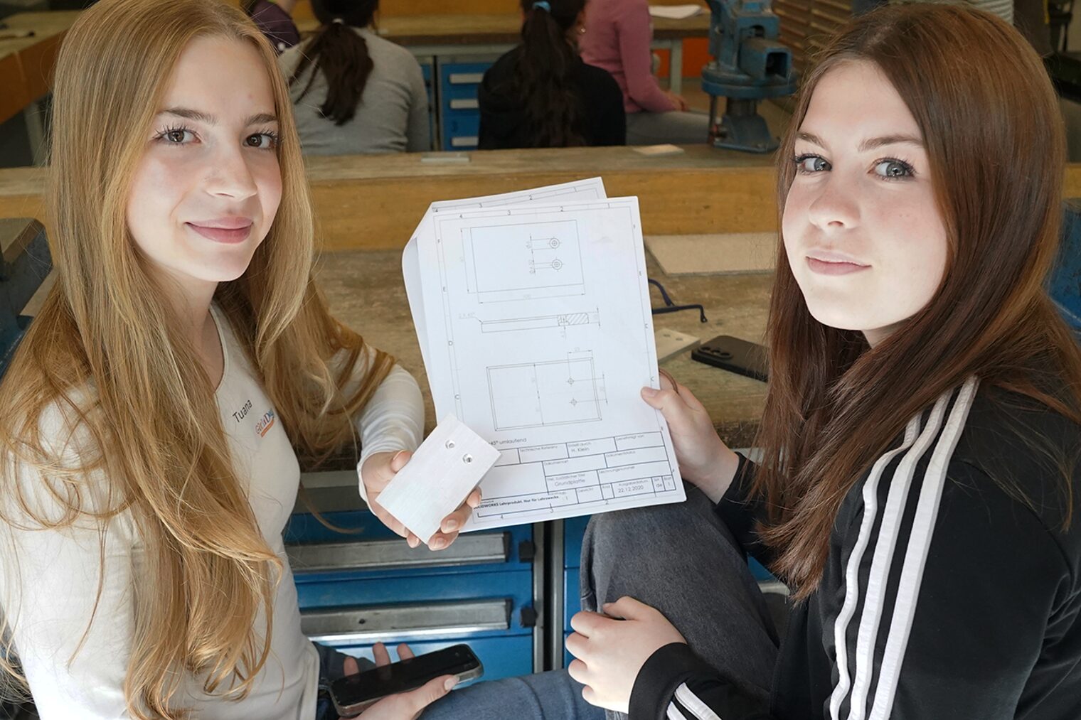 Zwei junge Frauen zeigen die Bauanleitung zu einem Handyhalter aus Metall und schauen in die Kamera.