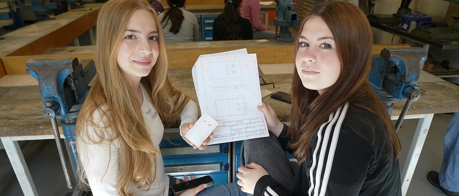 Zwei junge Frauen zeigen die Bauanleitung zu einem Handyhalter aus Metall und schauen in die Kamera.