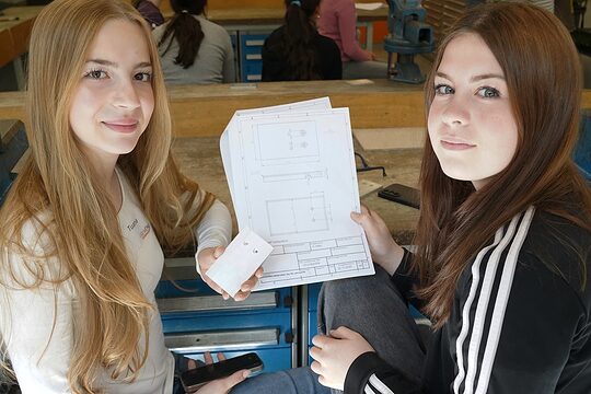 Zwei junge Frauen zeigen die Bauanleitung zu einem Handyhalter aus Metall und schauen in die Kamera.