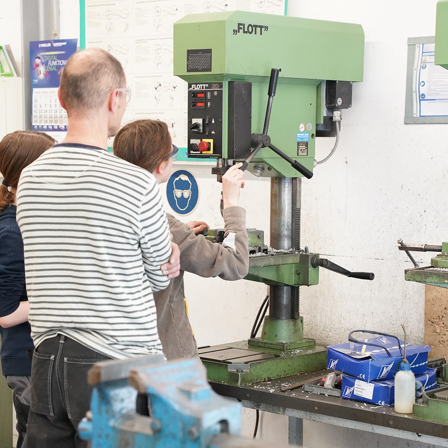 Mädchen stehen mit dem Ausbilder an der Bohrmaschine in der Metallwerkstatt.
