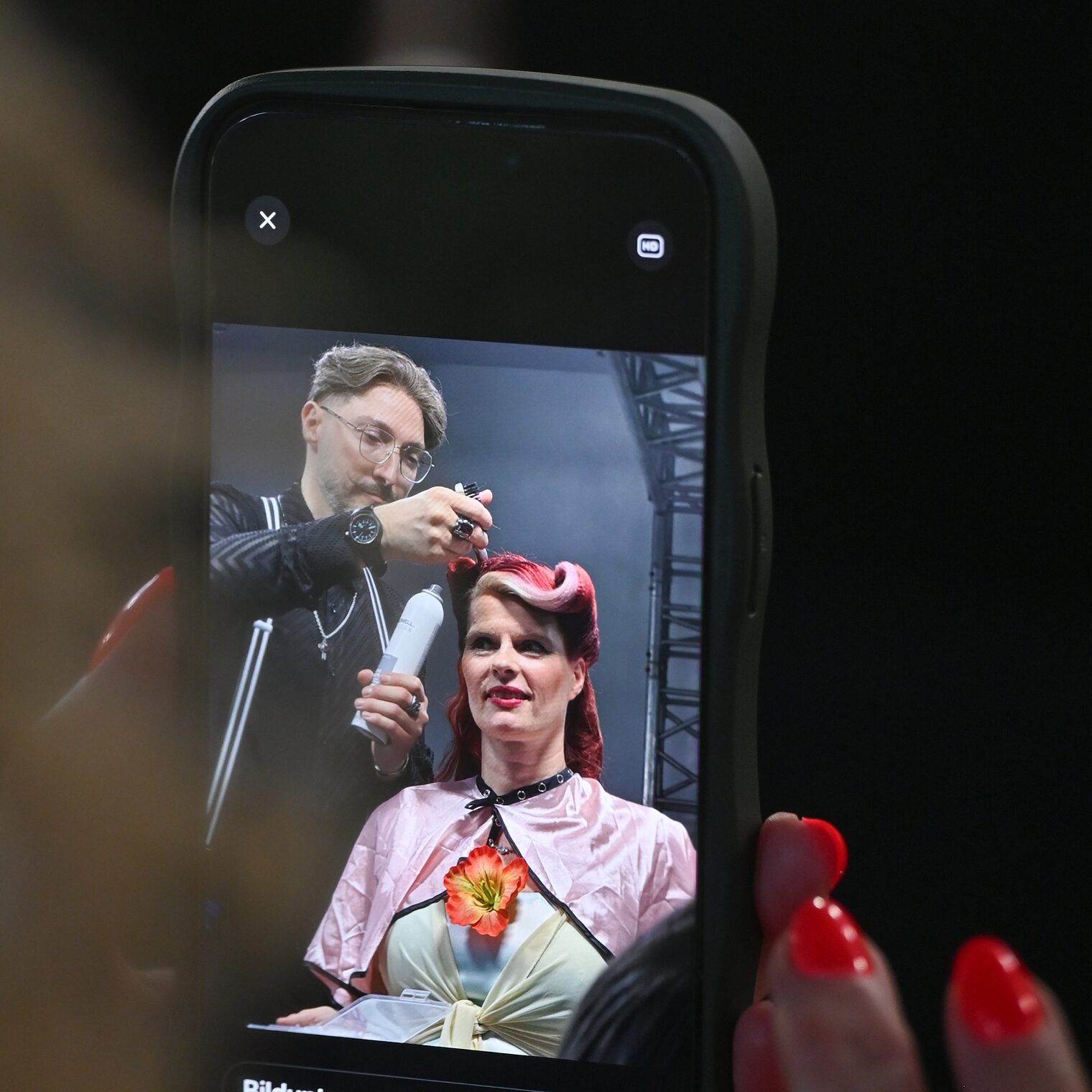 Ein Handybildschirm, auf dem ein Friseur mit seinem Model zu sehen sind, in den Händen einer Zuschauerin.