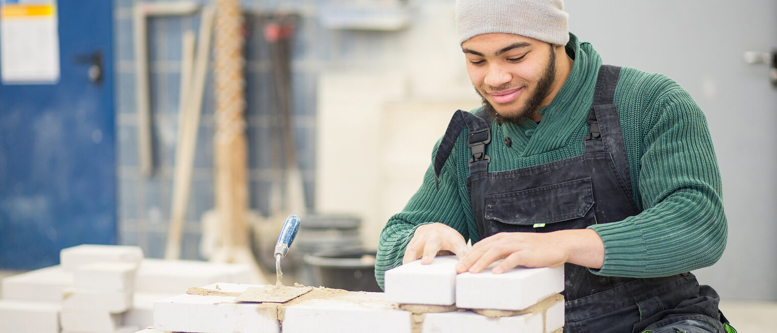Junger Mann in der Ausbildung als Mauerer setzt Steine mit Mörtel auf.