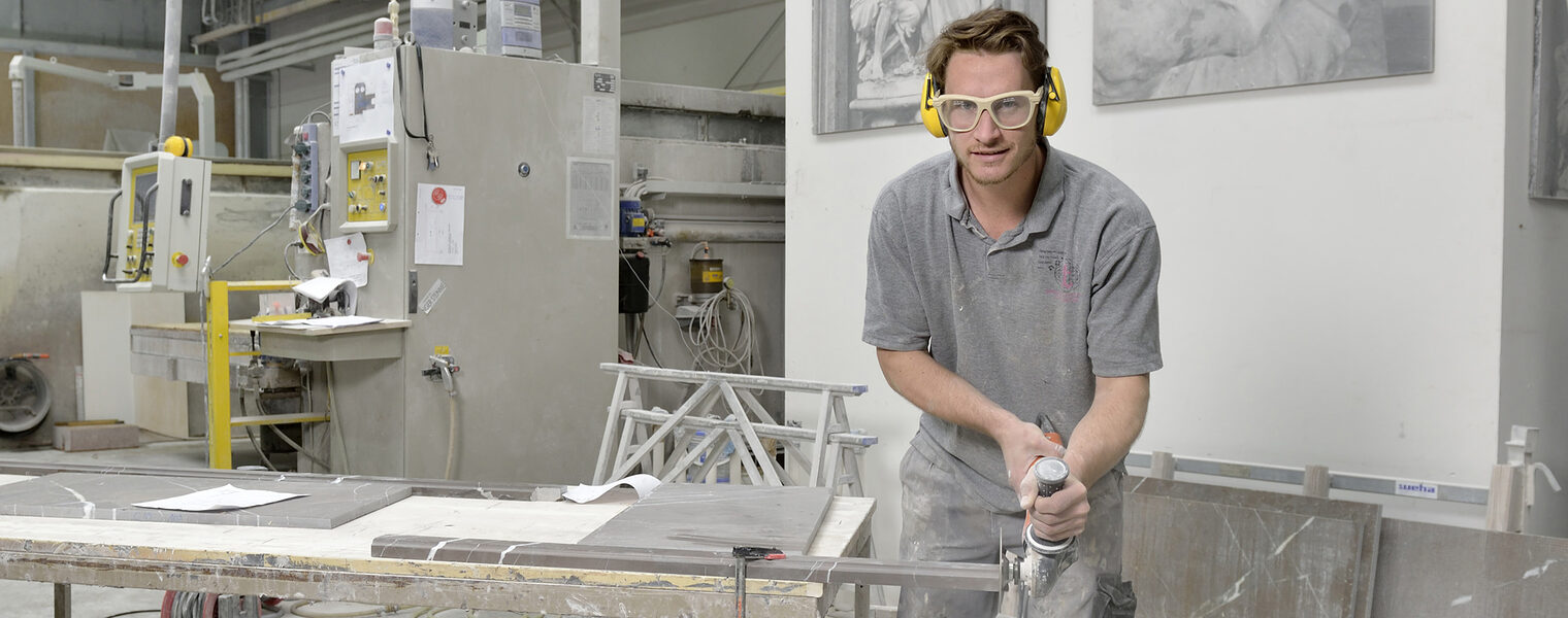 Gesamtsicht Werkstatt, Steinmetz fräst, Blick in die Kamera